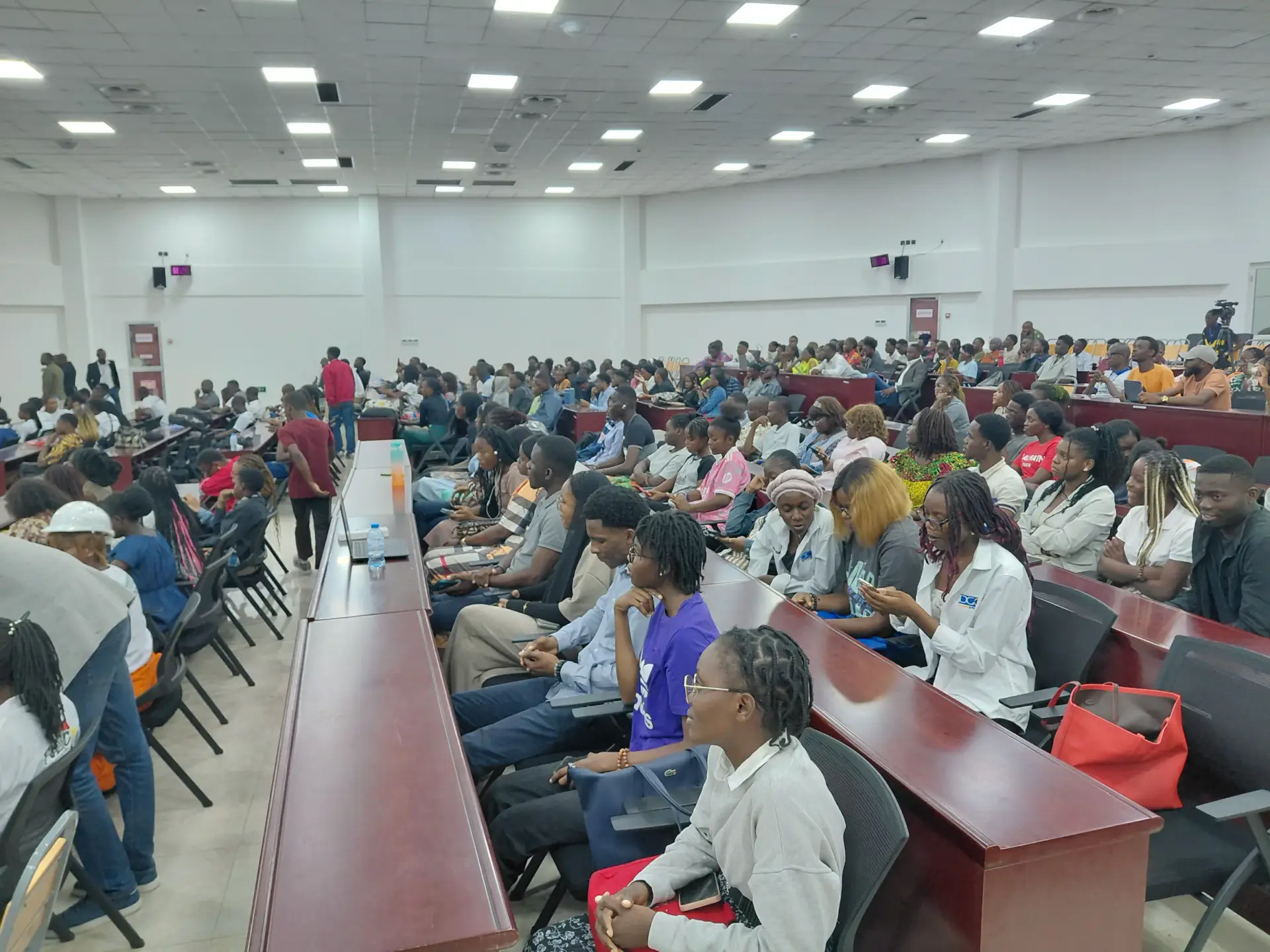 De nombreux étudiants, majoritairement jeunes, sont assis dans un grand auditoire. Certains écoutent une présentation, d'autres regardent leurs téléphones.